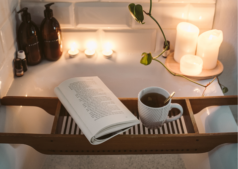 Una relajante escena de baño con un libro y un café, colocados en una bandeja de baño de madera. Las velas brillan agradablemente en el fondo.