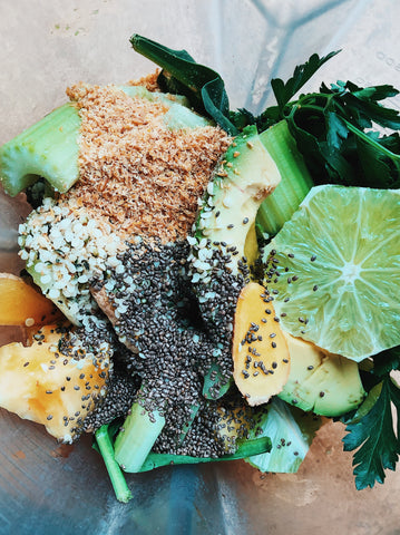 Ingredients for green smoothie all tossed into a blender: handfuls of spinach, parsley, ginger, lime, pineapple, avocado, stalks of celery, flax seed and chia seed.
