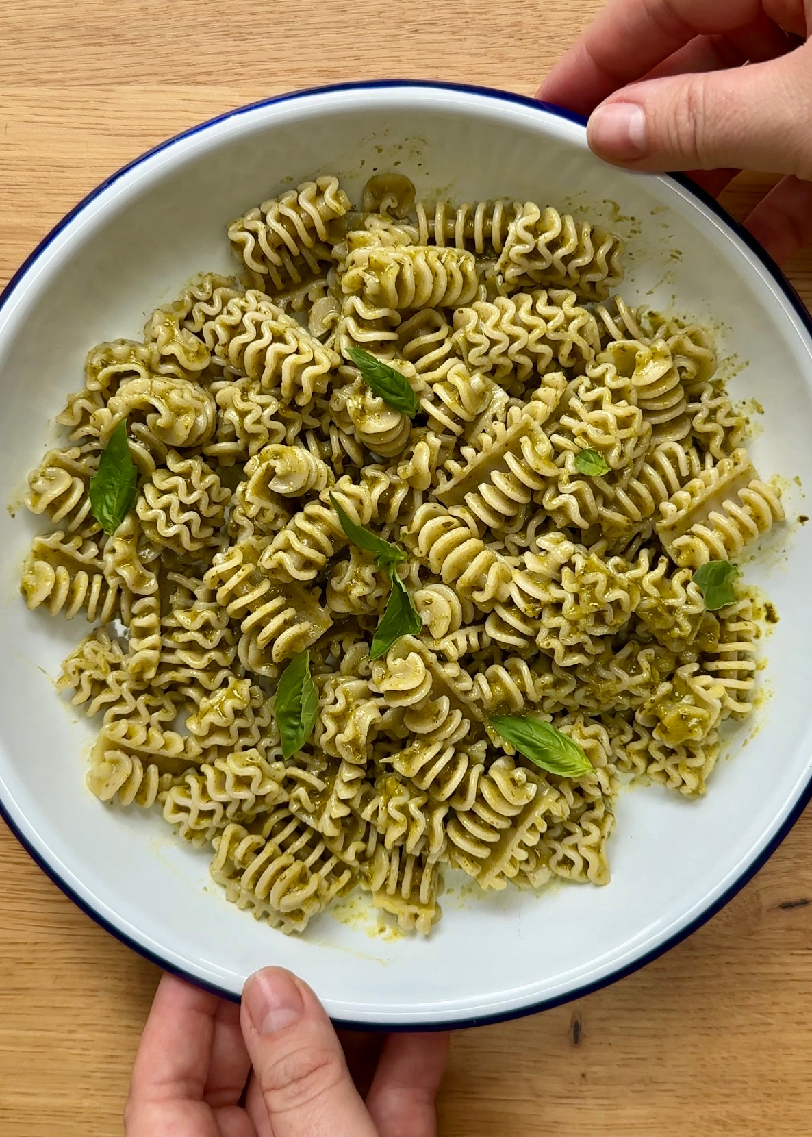 Close-up of fusilli pasta tossed in Saucerer's Chimichurri Pesto, garnished with fresh basil leaves in a white enamel bowl.
