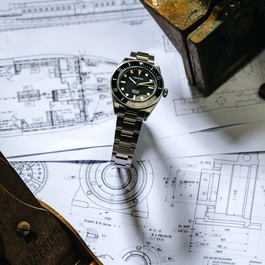 A wristwatch on engineering blueprints with a rusted metal object in the background.