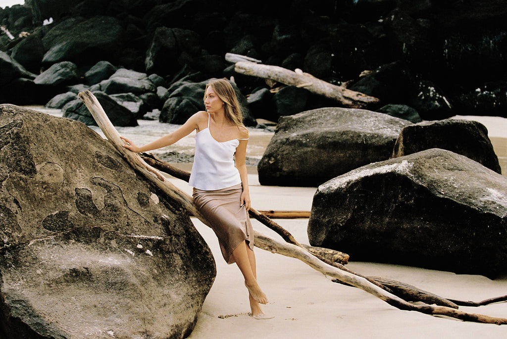 Model wearing white silk camisole and taupe silk skirt posing on a rock by the beach