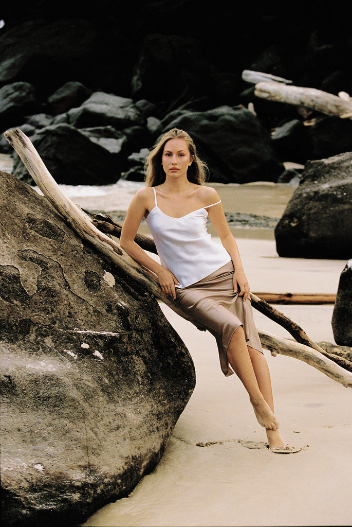 Woman wearing white silk camisole and taupe slip skirt posing on a rock by the beach