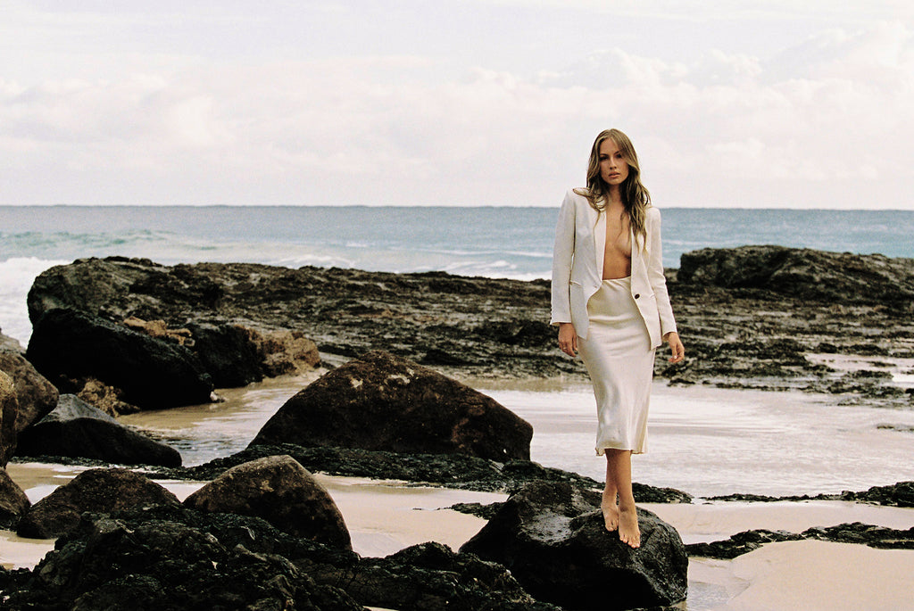 Model wearing white linen blazer and cream silk slip skirt posing on a rock by the beach