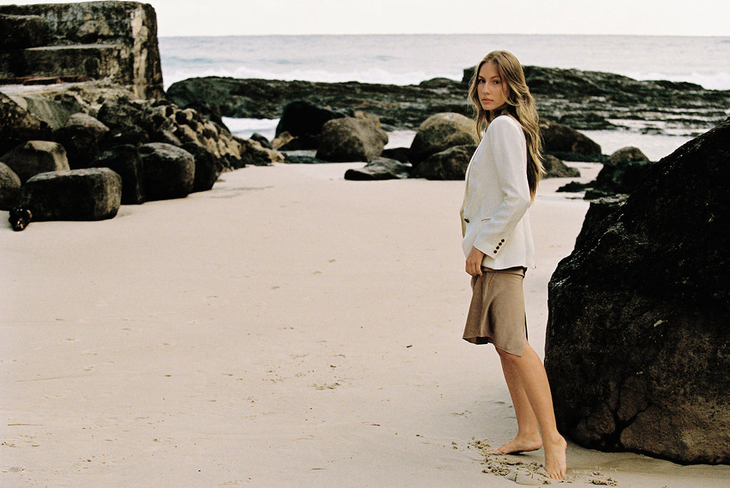 Model wearing white linen blazer and taupe slip skirt posing by the beach