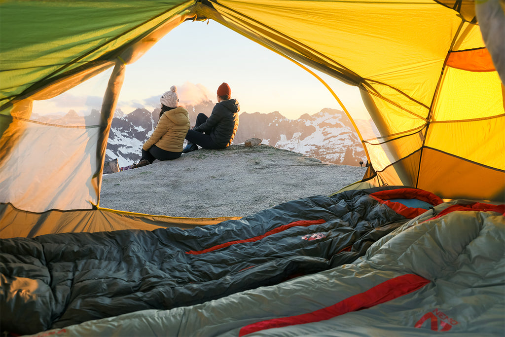 Man and woman sitting outside of a tent with two sleeping bags inside.