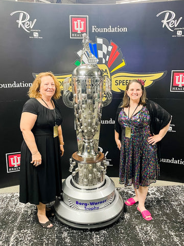 Two women in formal dresses agains a black backdrop with the Borg-Warner Indy 500 trophy between them