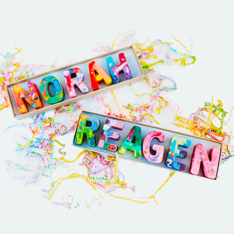 Personalized name crayons of NORAH and REAGEN in pastel rainbow colored crayon mix. Each letter has multicolor for the crayon. Boxed crayon set is on a white background with Easter grass around it as a photo prop.