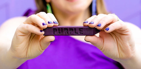 An Oversized purple crayon is being held that says the color name, PURPLE, engraved on the crayon
