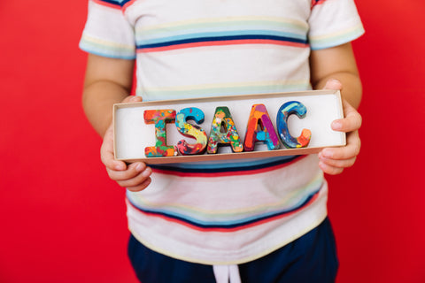 Image of a custom crayon name set that says ISAAC. A child is holding the rainbow colored letter crayons in a 8x2 gift box. The background of the photo is red. Personalized crayon gift for kids, rainbow crayon letters that spell out your name from crayon shop Art 2 the Extreme