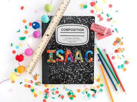 school supplies for kids, image is of a rainbow crayon name set that spells ISAAC on top of a black notebook with pencils, a ruler, and other supplies surrounding it.