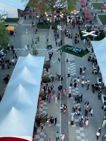 Image taken of the REV vip event at the Indianapolis Motor Speedway from above in the pergola. The tops of white tents are shown with people looking small like ants. There is a red and checkered carpet.