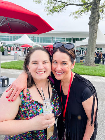 Two women hugging outside the Indianapolis Motor Speedway Rev red carpet event. One has a Simplicity cold-pressed cocktail beverage in hand with a checkered straw.