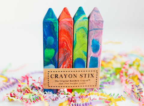 Colorful crayon Stix from crayon shop Art 2 the Extreme. These rectangular shaped, multicolored crayons come in a pack of four. Image has the shrink-wrapped rainbow crayon set showing with colorful crinkle paper and a white background as photo props.