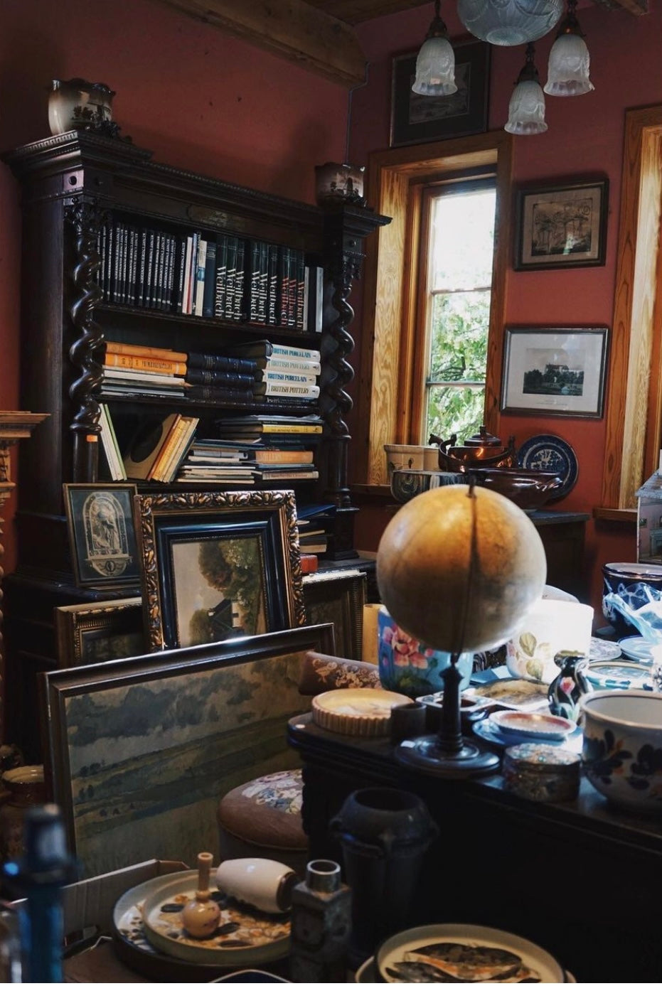 På bilden ser man ett gammalt stort skåp som är full med böcker. Framför den står ett antikt skrivbord med en glob på den. Hela rummet är full med saker.