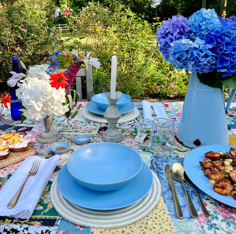 Blue ceramics on a colorful patchwork tablecloth with florals and a green garden background.