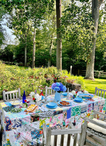 A colorful daytime table setting using blue ceramic dishes and a patchwork tablecloth. Green lawn and big trees are in the background.
