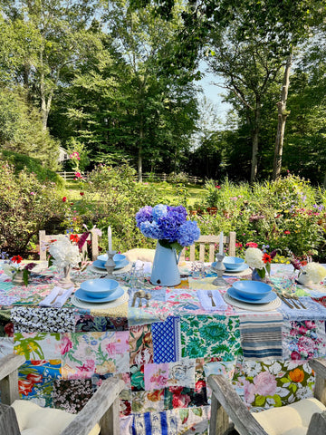 colorful summer table set for a daytime lunch. Blue ceramic dishes and blue hydrangeas are on the table. Greenery is in the background.