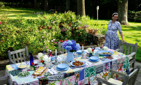 Meg is wearing a floral dress as she sets a summer lunch table with blue ceramics and a patchwork tablecloth.