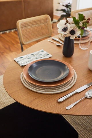 Ceramic plates, flatware, and linens set on a dining table with a bud vase and flowers.