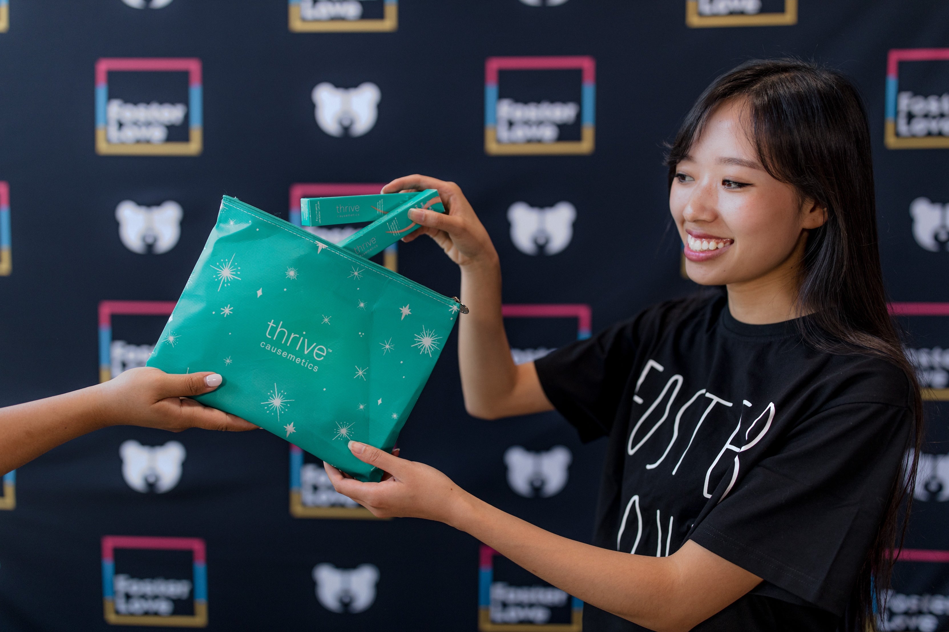 Woman smiling, receiving a teal cosmetic bag from another person, with a branded backdrop.