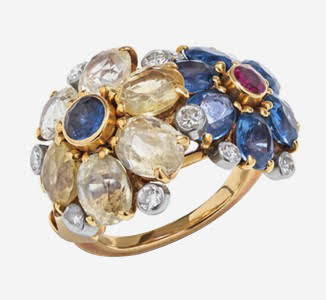 An overview photograph of a Cartier London ring made of diamonds and colored sapphires in a floral motif. One flower of yellow sapphire petals with a blue sapphire center and one flower of blue sapphire petals with a ruby center. Both flowers surrounded by round diamonds. All set in yellow gold with a yellow gold band. Photographed on a white background.
