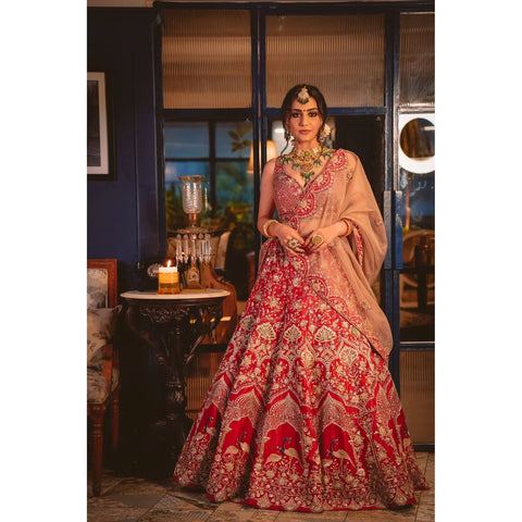 Deep red silk bridal lehenga choli with Mughal arches and peacock motifs in gold zardozi, resham and applique embroidery; paired with a beige gold tissue dupatta embroidered with floral zardozi buttis and a silk scallop border.