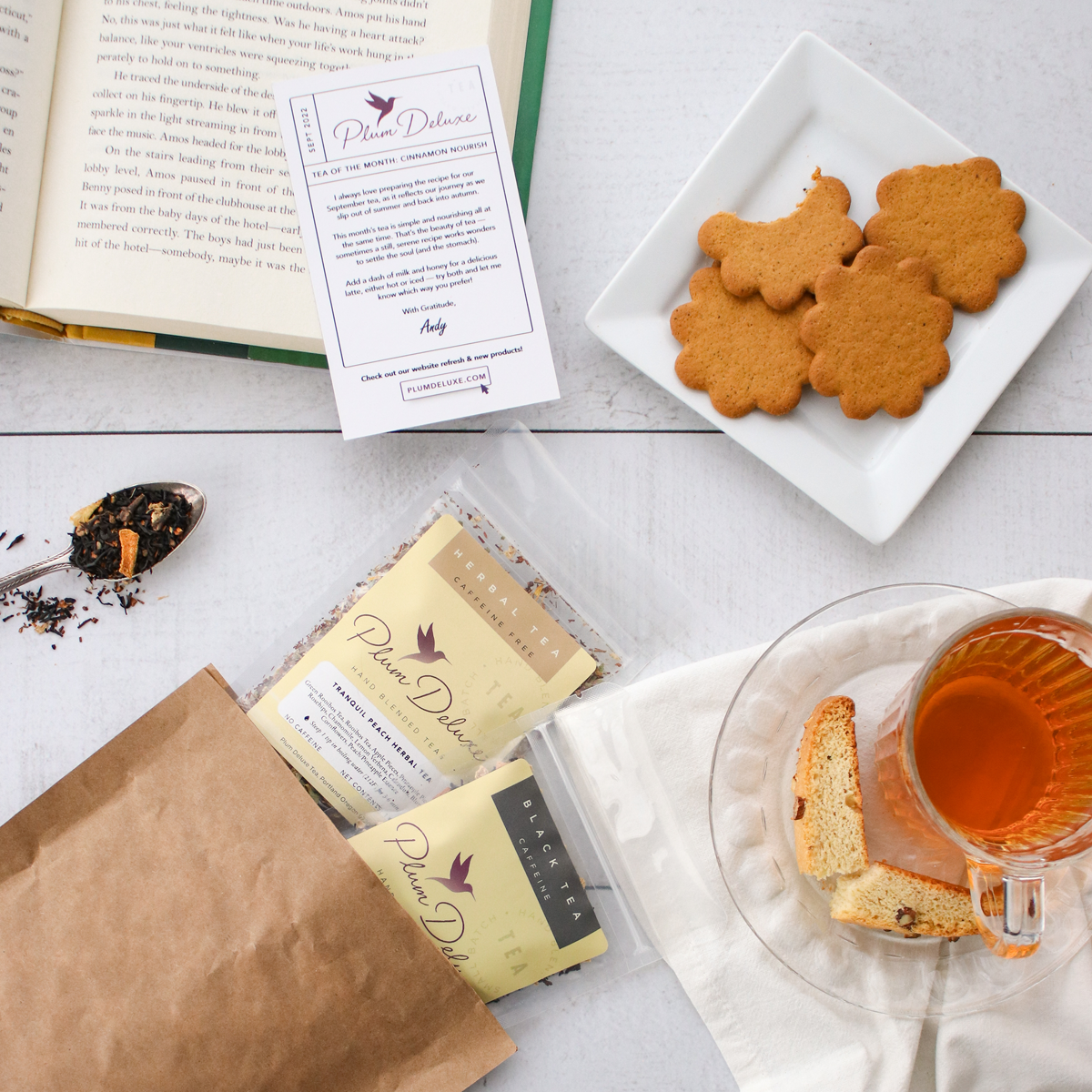 Overhead photo of a recyclable kraft paper envelope with two packages of tea sticking out. Next to it on a white wooden table is a glass teacup and saucer, a small plate of cookies, and an open book.