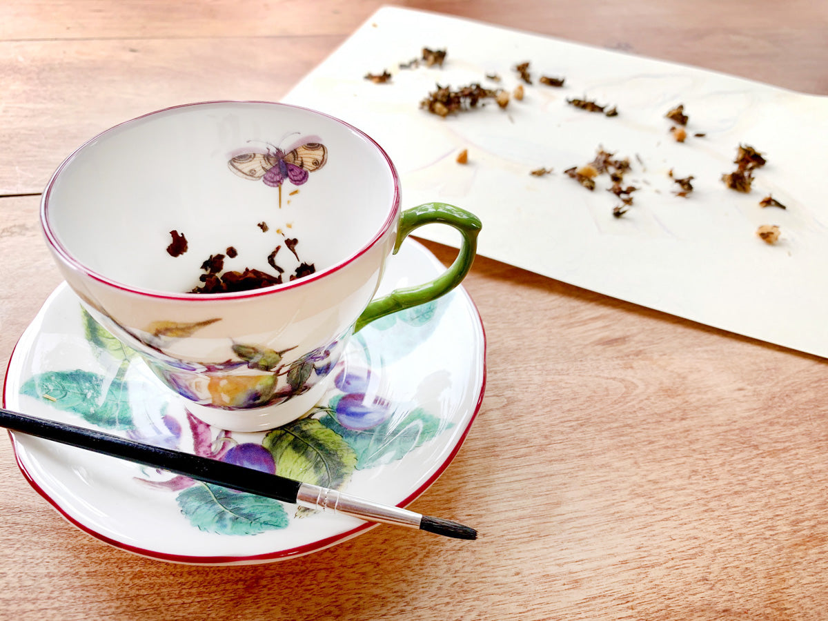 Photo of a teacup and saucer next to a sheet of paper on a wooden table. A paintbrush rests on the saucer.