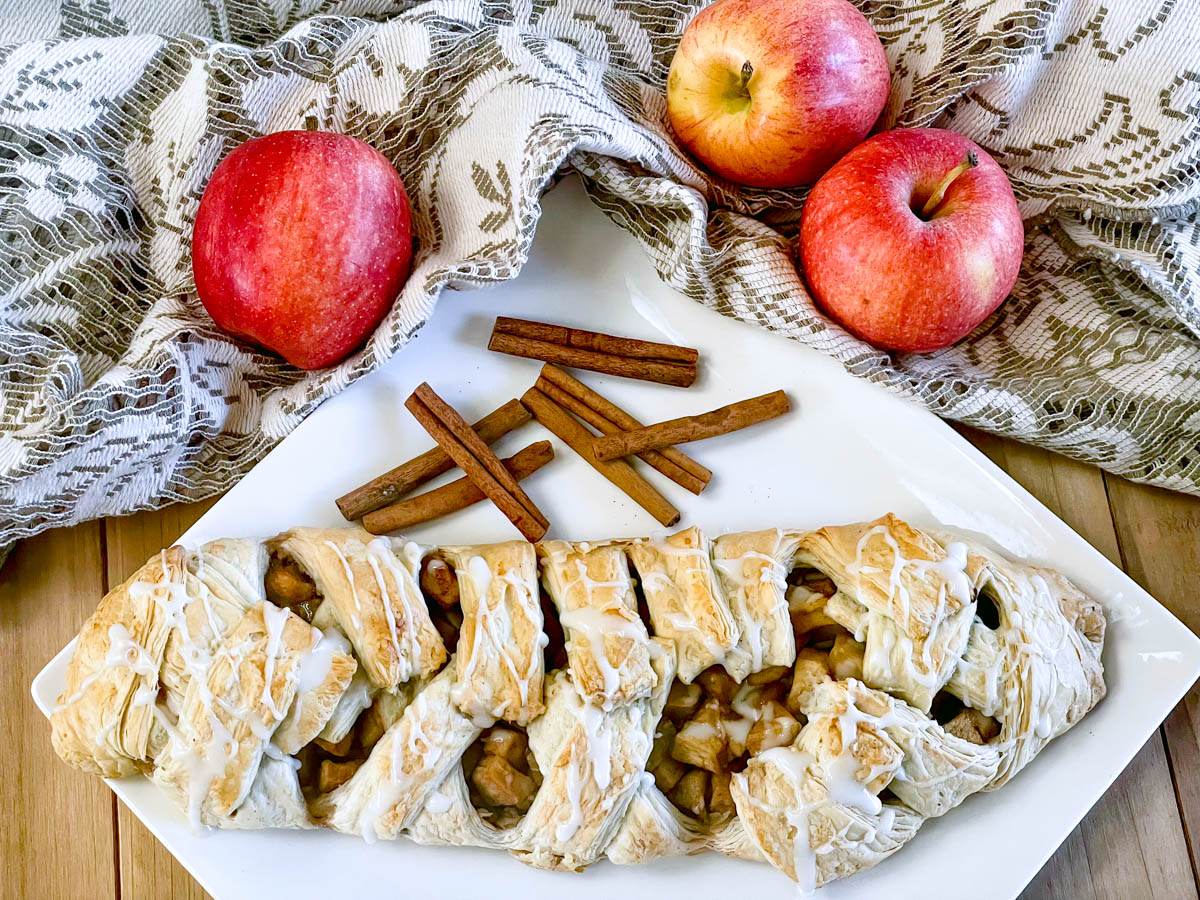 A whole apple strudel sits on a plate with cinnamon sticks and fresh apples. The golden filling is visible through the slits on top.