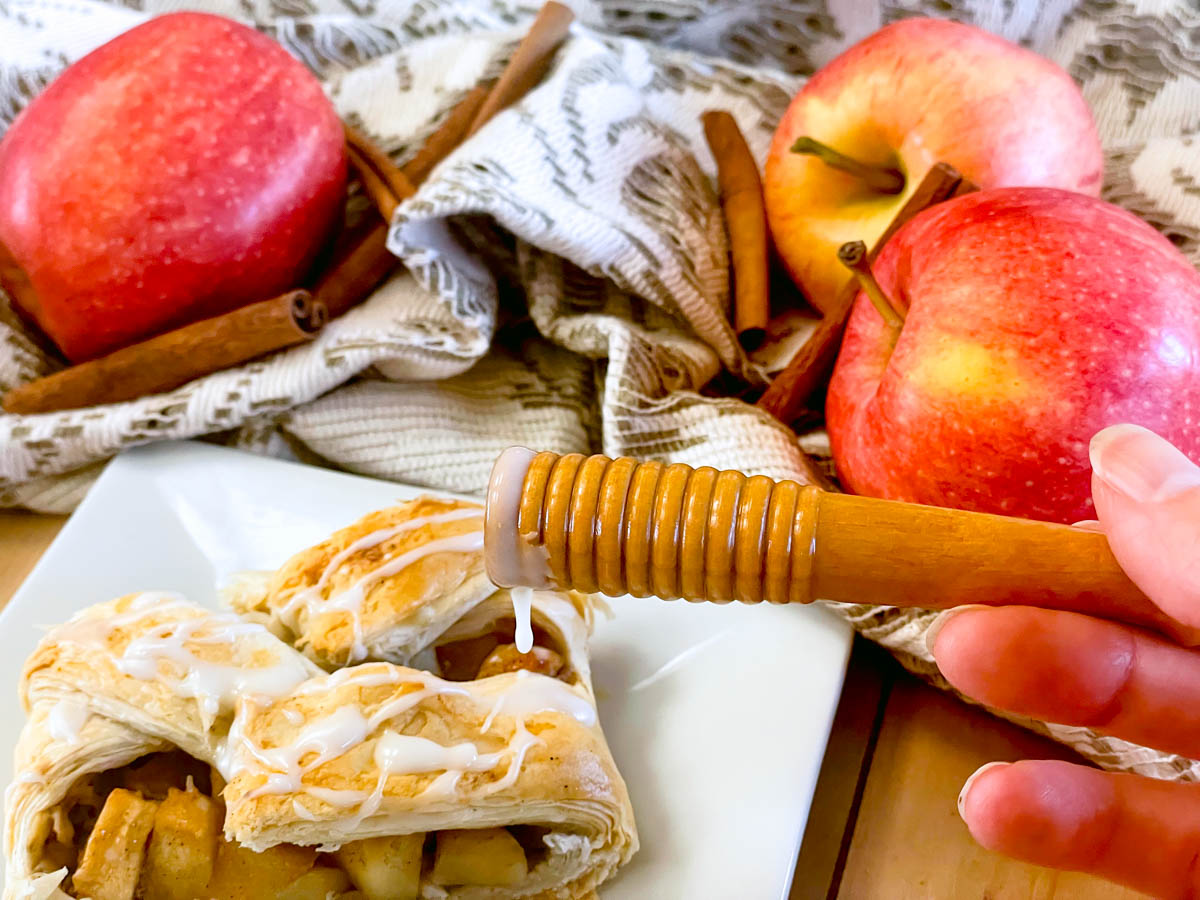 Drizzling icing onto a freshly baked apple strudel surrounded by fresh apples and cinnamon sticks.