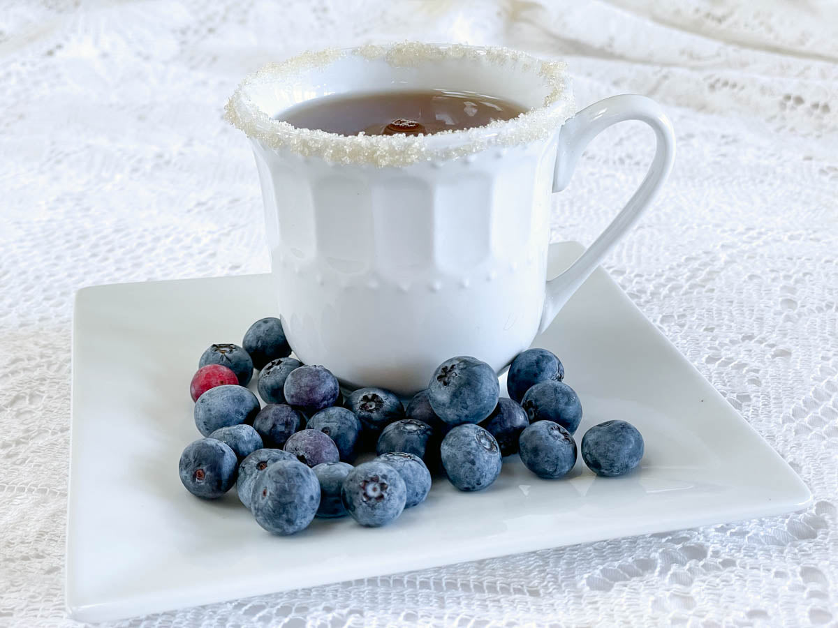 A cup of tea with a rim of sugar sits on a saucer surrounded by fresh blueberries.