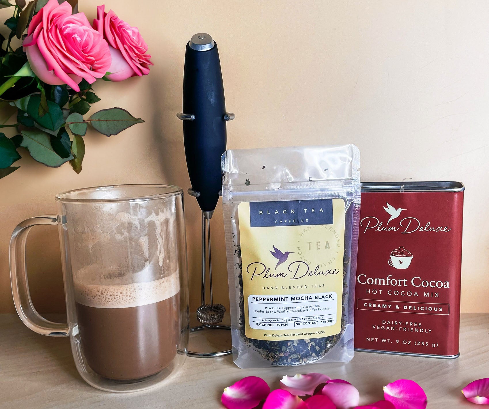 A glass mug of tea-infused cocoa and frothed oat milk sits on a counter next to a Latte Whisk, a bag of Peppermint Mocha loose tea, and a tin of Comfort Cocoa Mix. Pink roses can be seen in the background, and pink rose petals are scattered on the table.