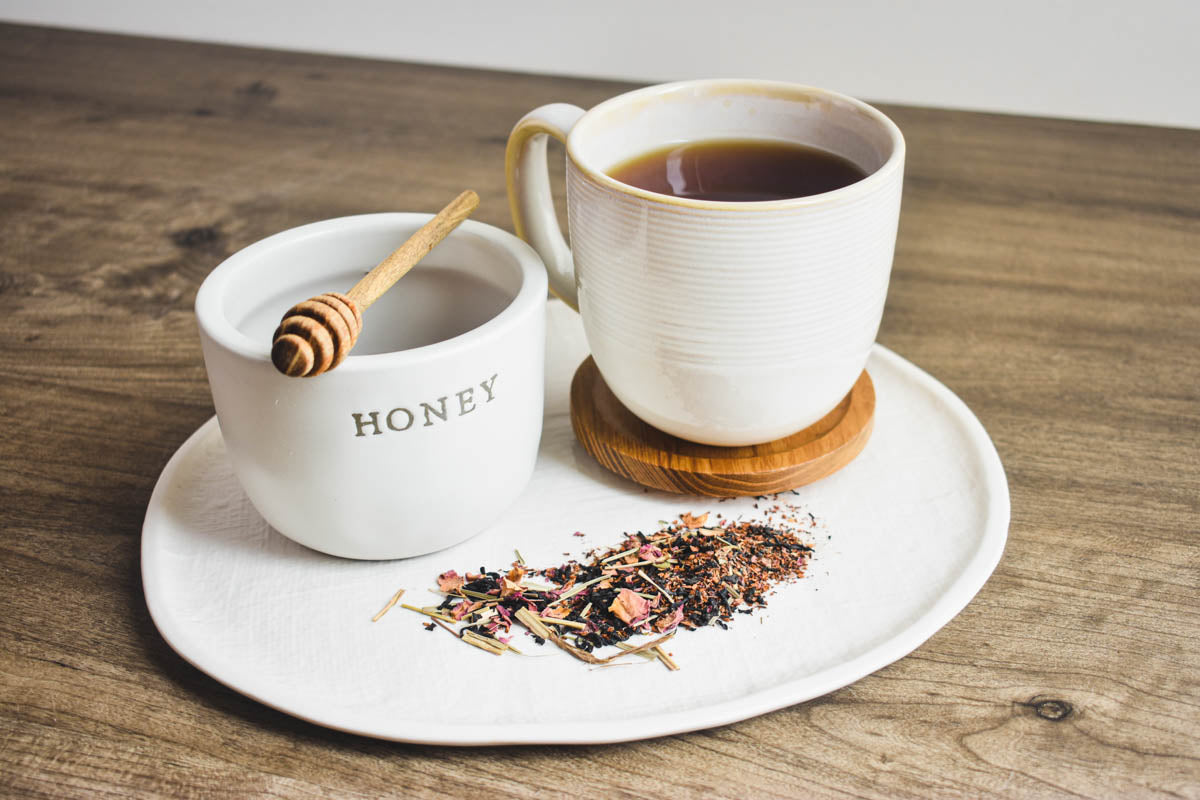 Mugs of honey and black tea sit together on a large plate with loose-leaf black tea.
