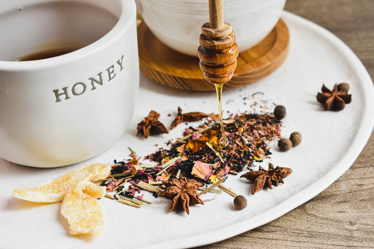 Honey is being drizzled onto a mixture of loose-leaf black tea and chai spices, notably ginger, allspice and star anise.