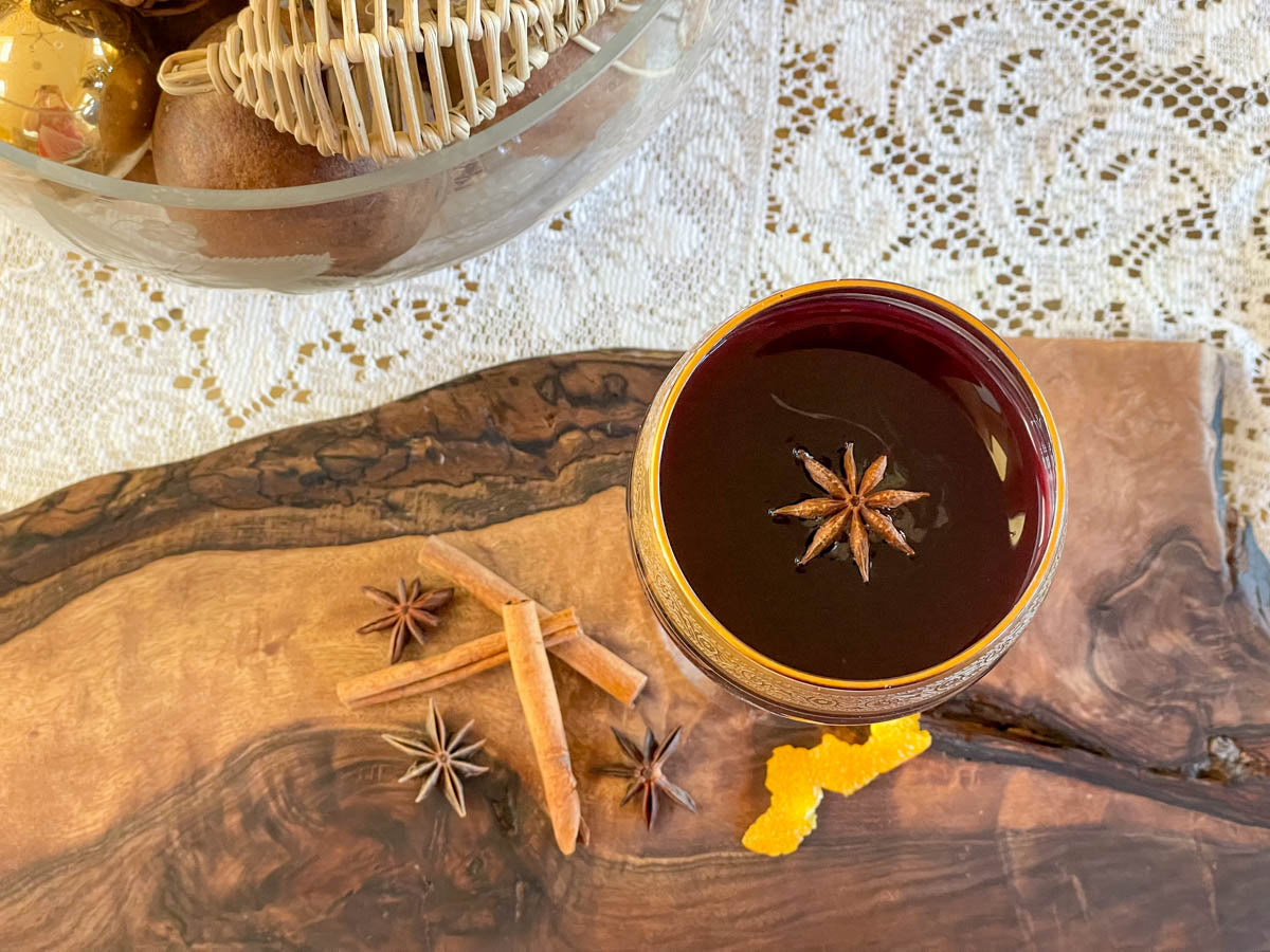 A glass of wine with a star anise pod floating in the glass sits on a wooden surface with an orange rind off to the side. The table is decorated with a lace runner.