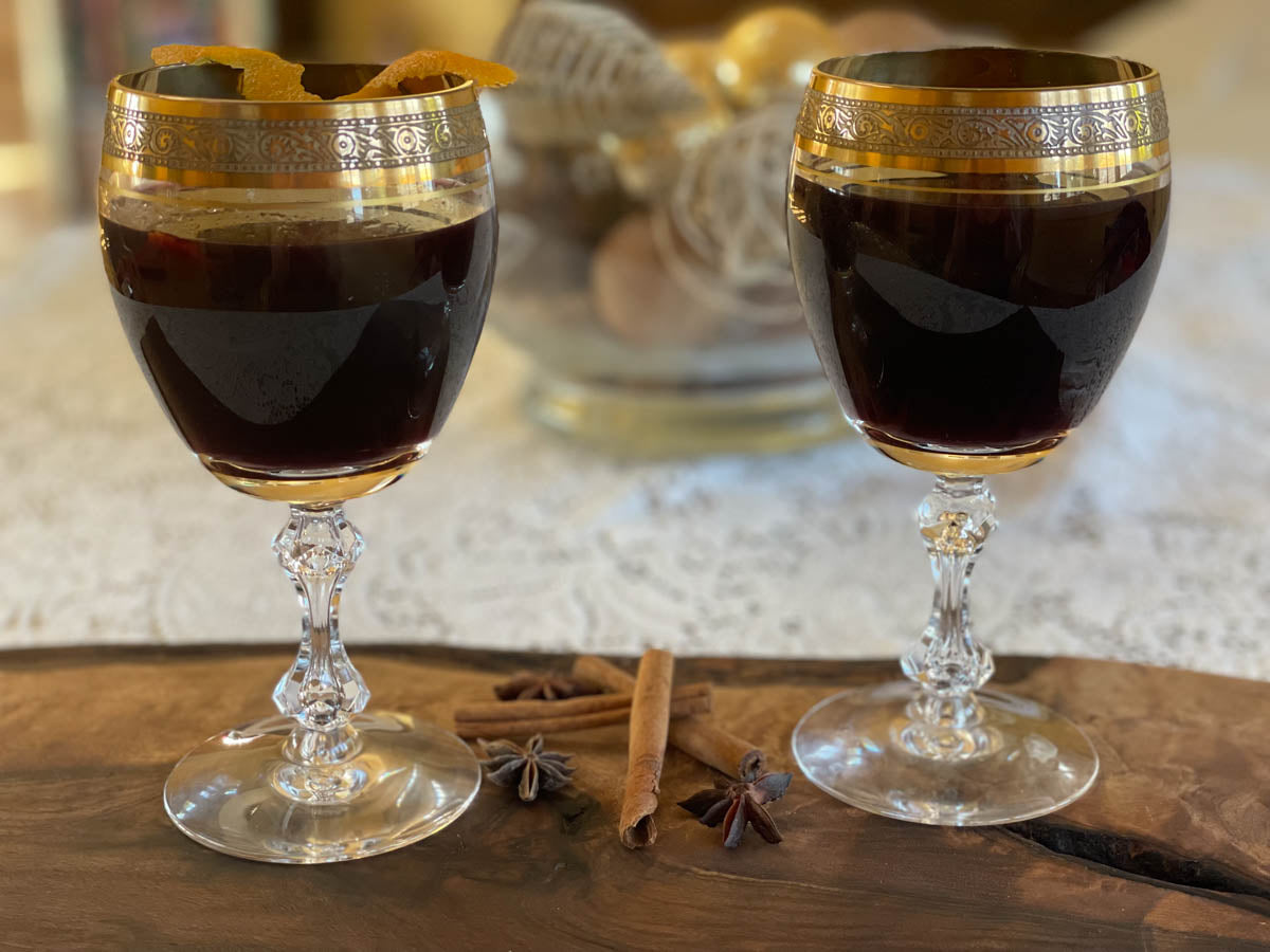 Two gold-rimmed glasses of mulled wine sit on a wooden table in front of a lace table runner.