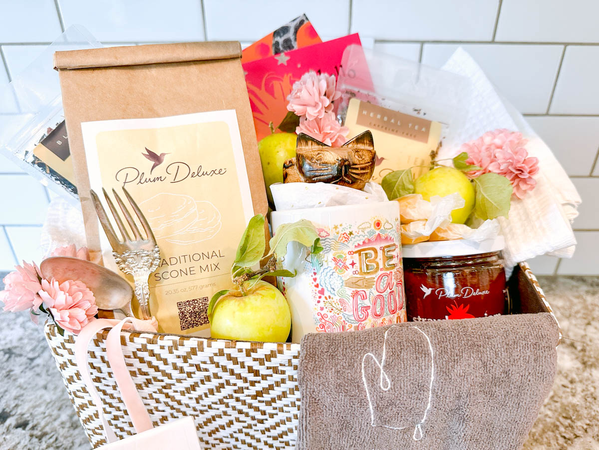 A large tea gift basket stuffed full of goods such as fresh fruit, scone mix, tea towels and hand-written cards.