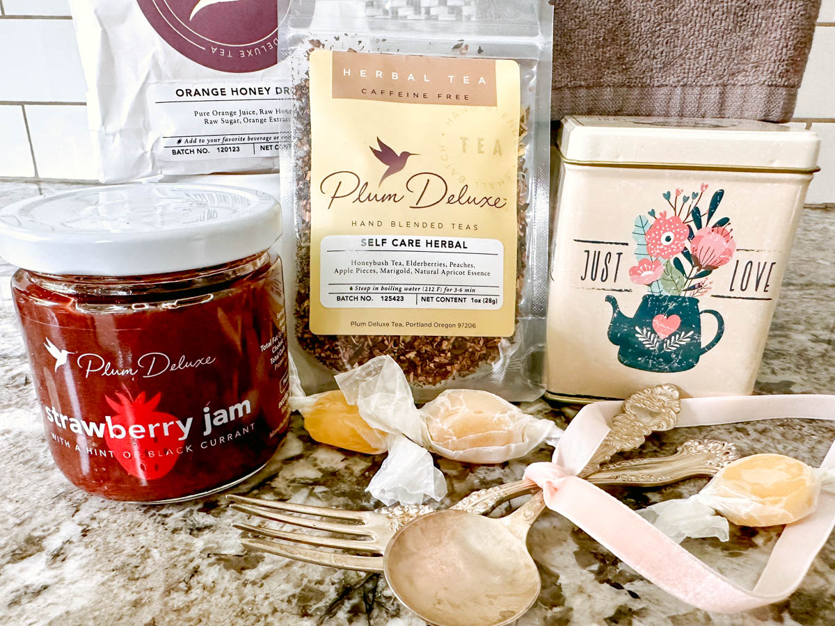 Assorted things to include in a tea gift basket, including loose-leaf tea, a tea tin, strawberry jam and more.