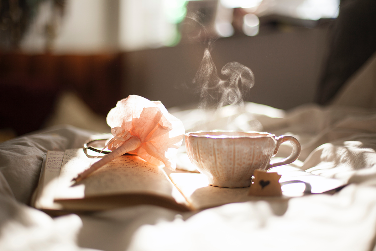 A steaming cup of tea and a pen with a pink flower on it sit on the pages of an open journal.