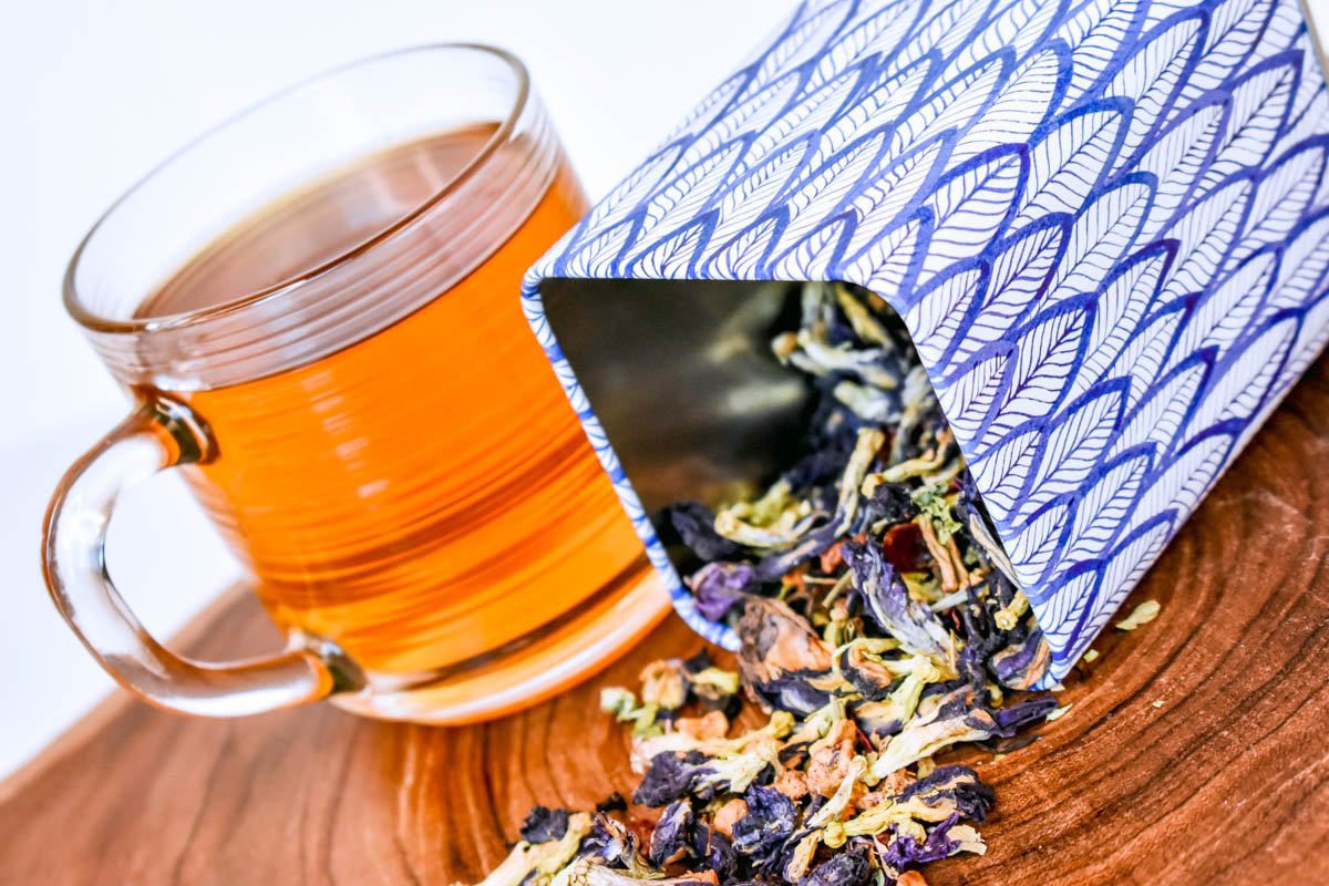 A glass of tea sits next to a tea tin from which spills out Magical Butterfly loose-leaf tea.