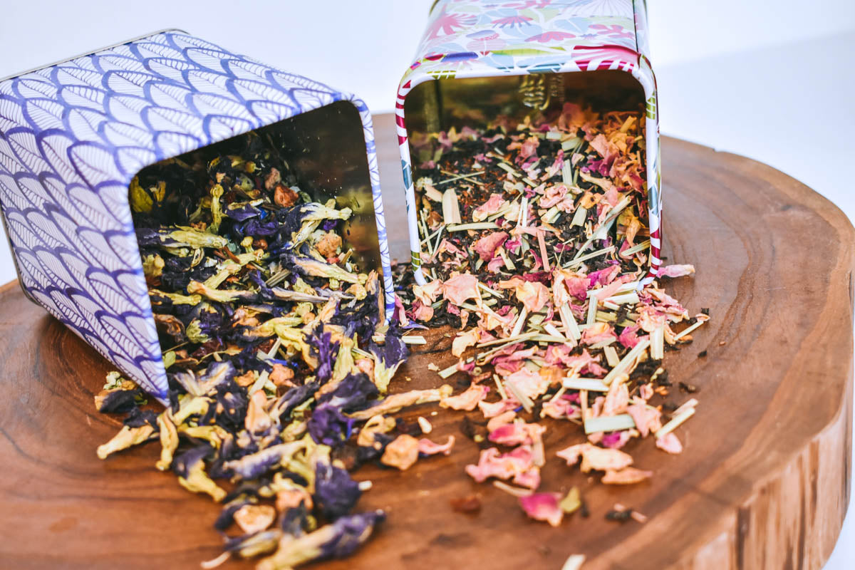 Magical Butterfly herbal tea and Portland Rose City chai sprawl out from their matching colored tea tins onto a wooden surface.