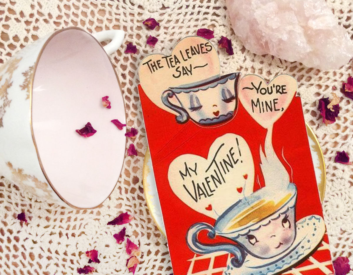 Overhead view of a vintage valentine card, a rose quartz, a white and gold teacup on its side, and scattered rose petals on a lace tablecloth.