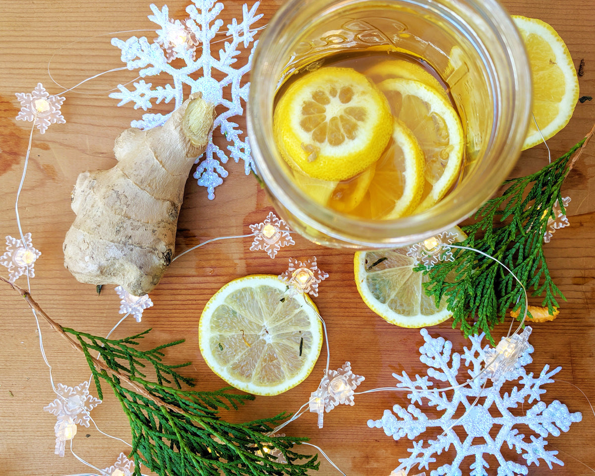 Overhead view of a jar full of honey, fresh ginger, and lemon slices on a wooden board. It is surrounded by fresh ginger root, lemon slices, evergreen boughs, decorative snowflakes, and twinkle lights.
