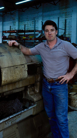 An image of Mack proudly standing next to an automatic screw machine used in crafting buckles for Mack belts. Mack, with a confident stance, showcases the manufacturing process. The machine's intricate details and Mack's presence convey the craftsmanship and dedication behind each buckle's creation, offering insight into the meticulous production of Mack belts.