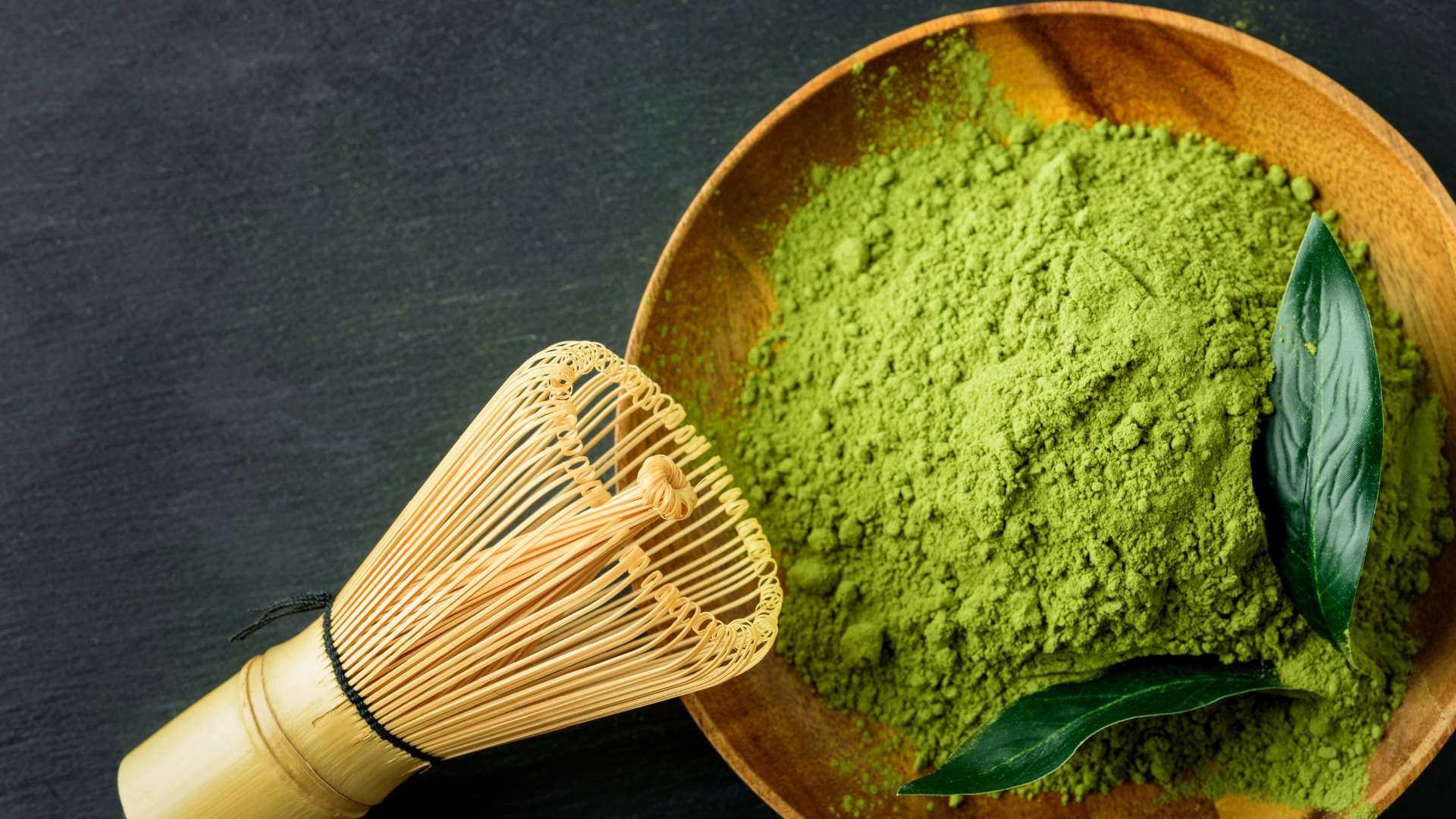 Matcha green tea powder in a bowl next to a bamboo matcha whisk