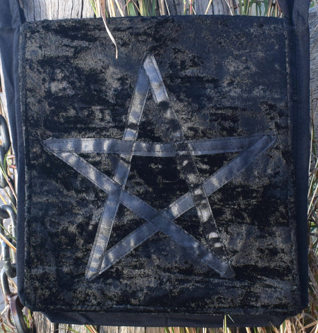 A black shoulder bag with a satin ribbon pentagram sewn on the front resting on weathered timber