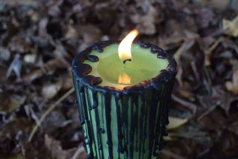 A green candle with black drips with its wick alight rests on a bed of autumn leaves.