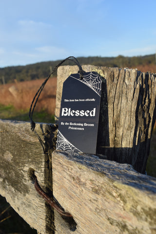 A black tag with string saying "this item has been officially blessed by the Beckoning Broom Priestesses" on it rests on a rock.