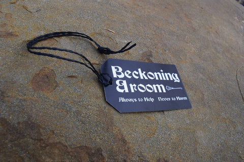 A black tag with string saying "Beckoning Broom Always to Help Never to Harm" on it rests on a rock.