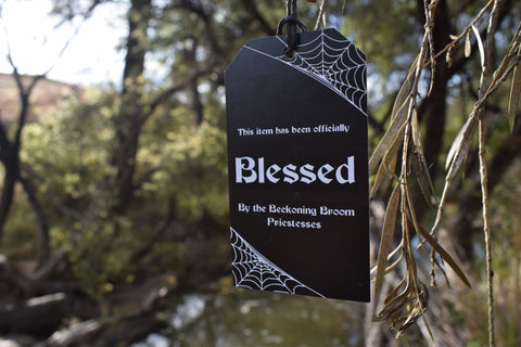 A black tag with string saying "this item has been officially blessed by the Beckoning Broom Priestesses" on it hangs from a branch.